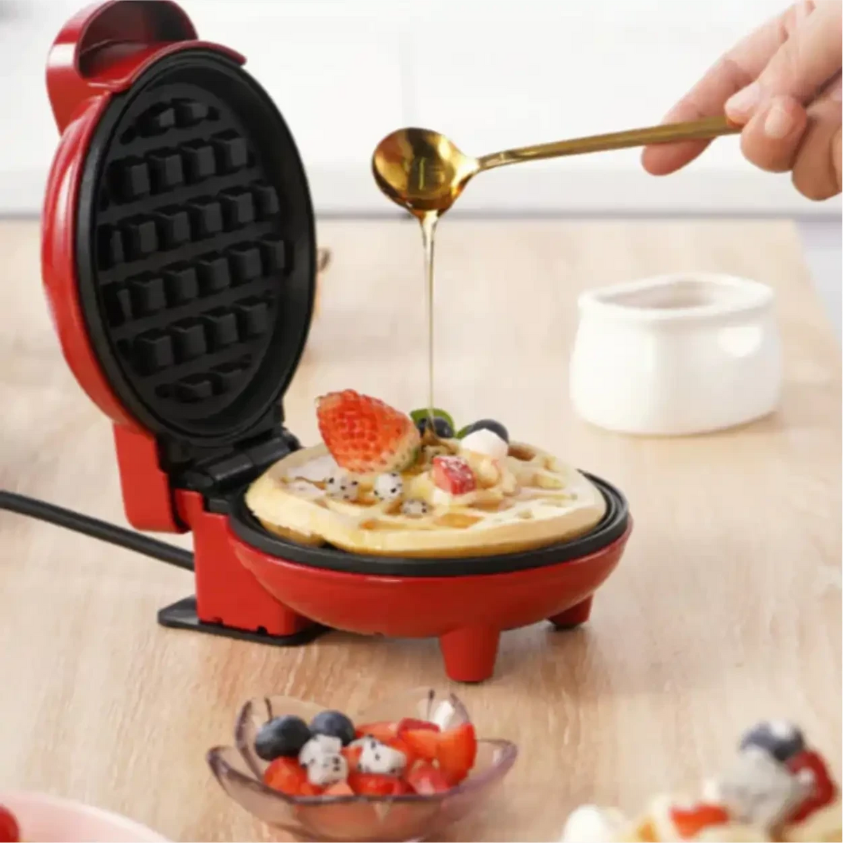 Máquina de waffle vermelha compacta Decoricasa com waffle fresco sendo coberto por mel e frutas em uma bancada
