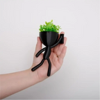 Mini vaso alpinista Decoricasa preto em formato de boneco segurando planta artificial verde na palma da mão
